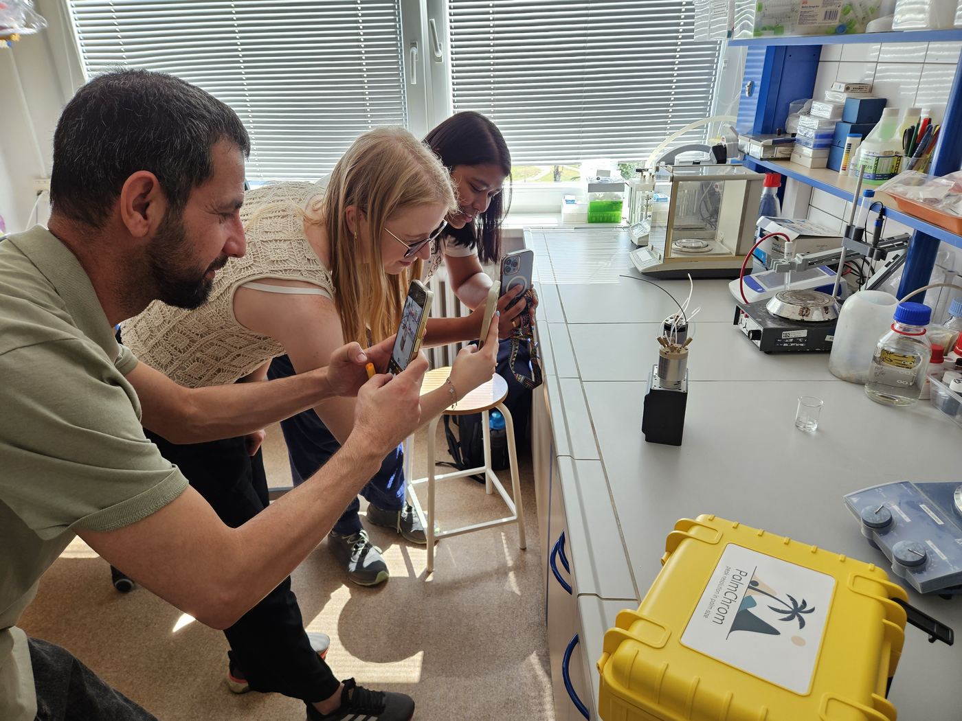 Three researchers photographing the PalmChrom prototype on a laboratory bench with their phones, the yellow PalmChrom case in the foreground