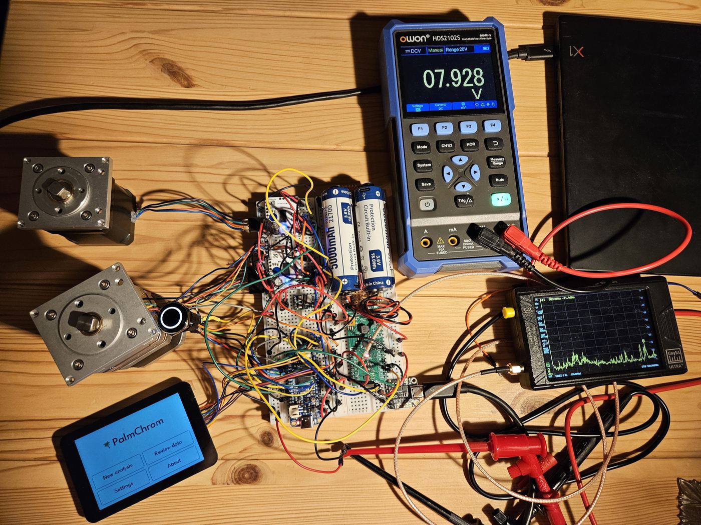 Breadboard electronics, batteries, two stepper motors and the touchscreen spread over a wooden table, next to a handheld oscilloscope and a tinySA spectrum analyzer
